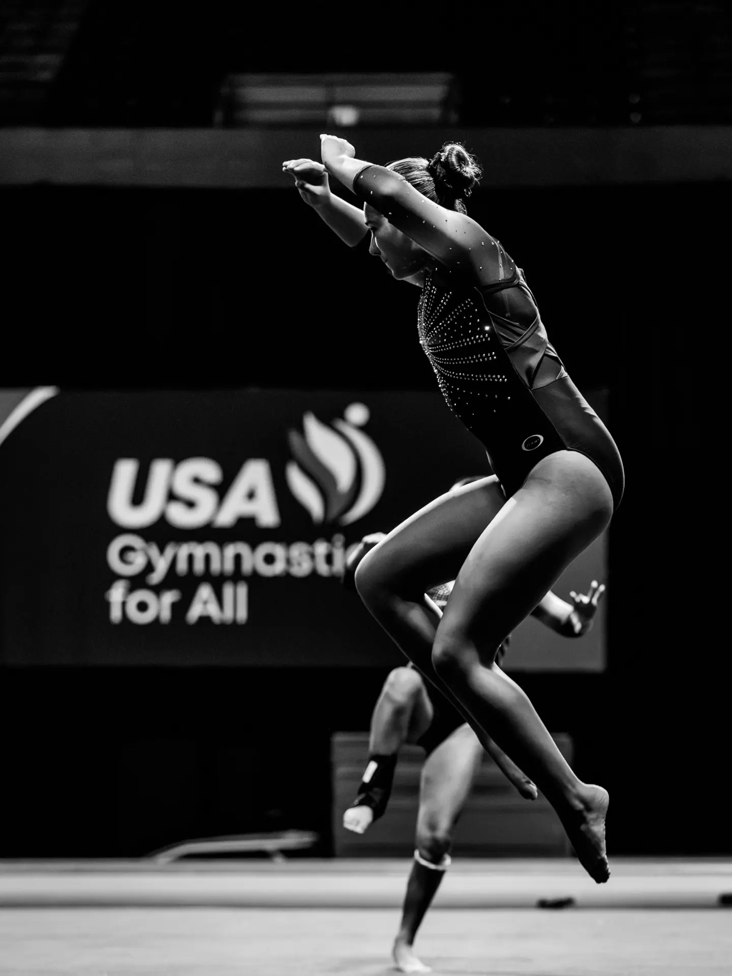 A performer beside a USA Gymnastics for All banner, black and white